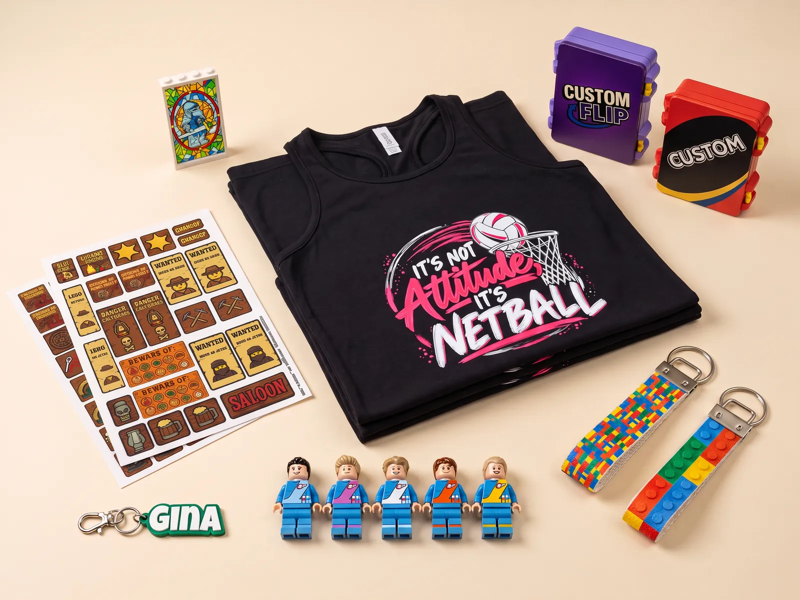 a flat-lay of recent jobs: printed netball singlets, sticker sheets, two game cases, a stained-glass LEGO panel, five minifigures, a name keyring and key straps